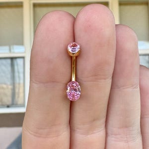 May include: A gold-toned curved barbell body jewelry piece featuring two pink faceted stones. The top stone is round and smaller, while the bottom stone is oval-shaped and larger. The metal bar connects the two stones in a vertical orientation.