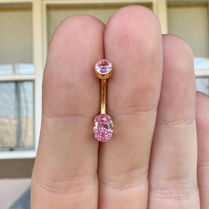 May include: A gold-toned curved barbell body jewelry piece featuring two pink faceted stones. The top stone is round and smaller, while the bottom stone is oval-shaped and larger. The metal bar connects the two stones in a vertical orientation.