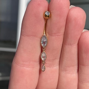 Puede incluir: Anillo de ombligo dorado con una sola piedra redonda transparente en la parte superior y tres piedras transparentes en forma de marquesa que cuelgan.
