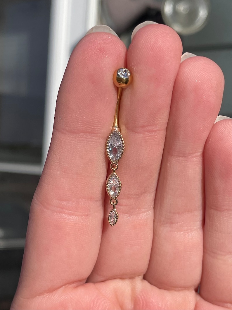 Puede incluir: Anillo de ombligo dorado con una sola piedra redonda transparente en la parte superior y tres piedras transparentes en forma de marquesa que cuelgan.