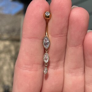 Puede incluir: Anillo de ombligo dorado con una cadena colgante de tres piedras transparentes en forma de marquesa.