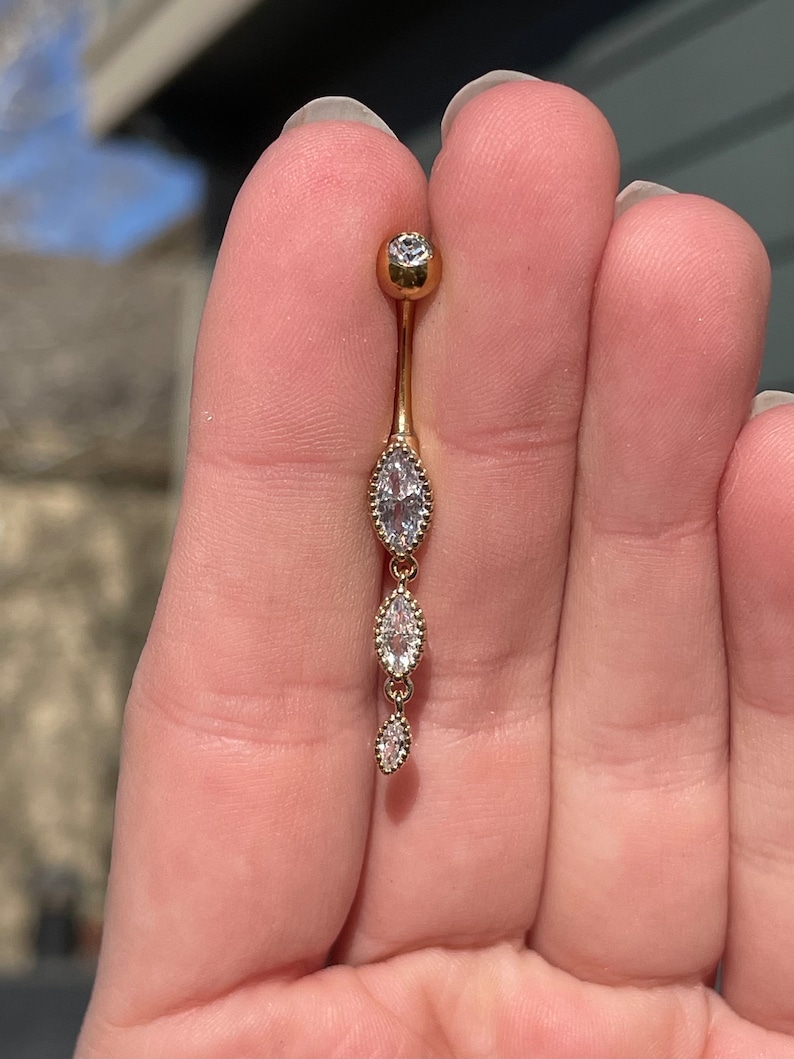 Puede incluir: Anillo de ombligo dorado con una cadena colgante de tres piedras transparentes en forma de marquesa.