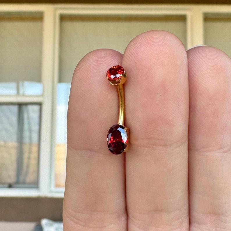 May include: A gold-toned curved barbell navel ring featuring two faceted, deep red stones. The smaller round stone is set at the top, while a larger oval-shaped stone is positioned at the bottom, both secured in polished metal settings.