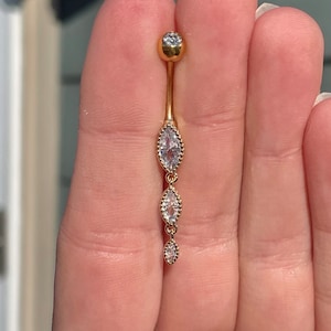 Puede incluir: Anillo de ombligo dorado con una cadena colgante de tres piedras claras en forma de marquesa.