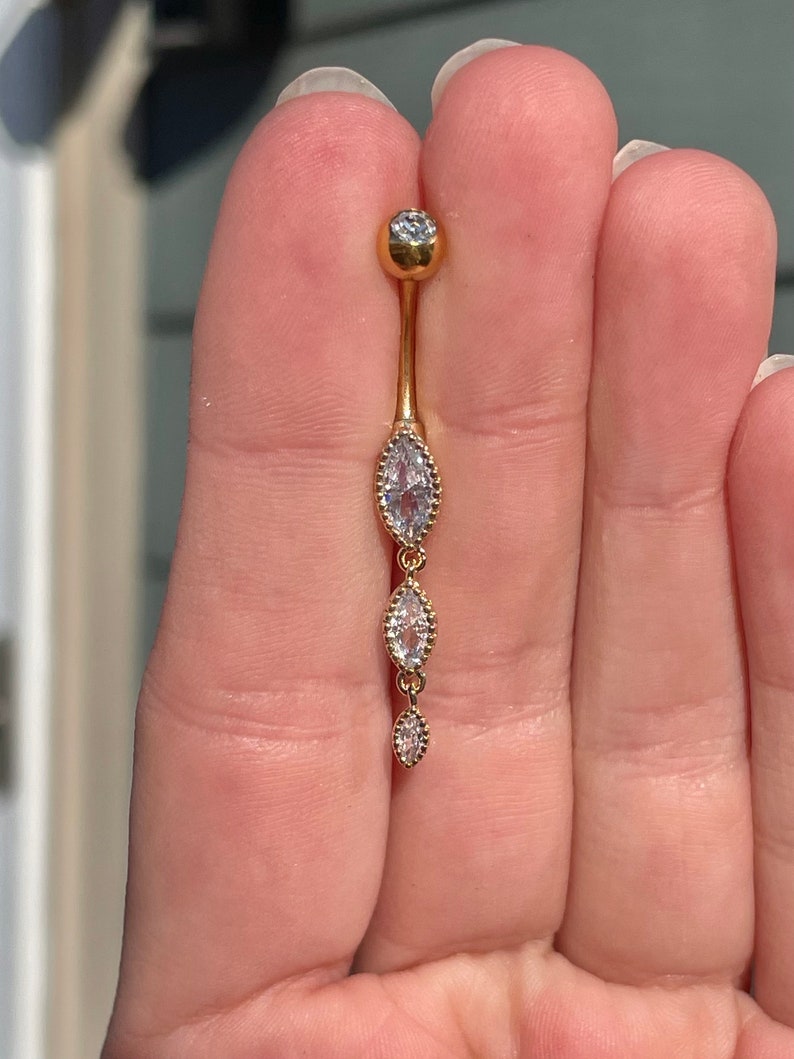 Puede incluir: Anillo de ombligo dorado con una cadena colgante de tres piedras claras en forma de marquesa.