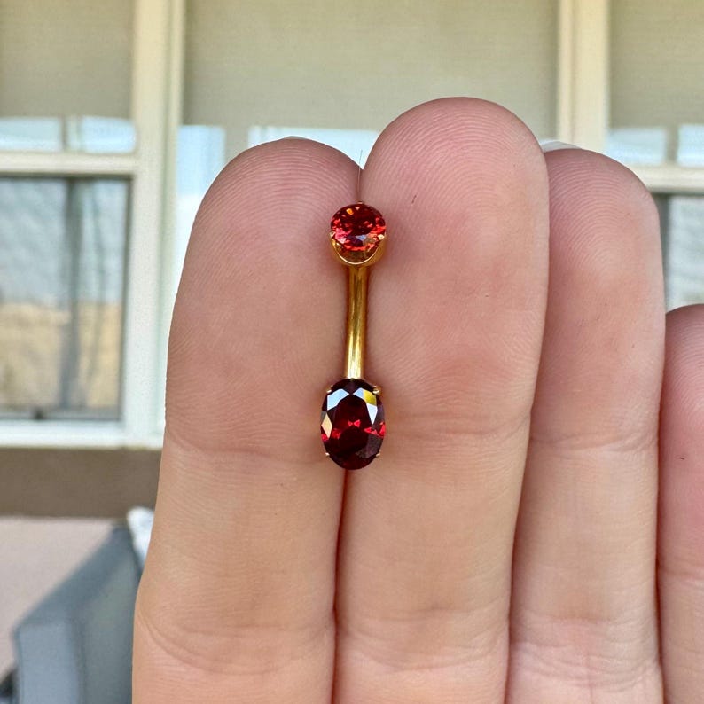 May include: A gold-toned curved barbell body jewelry piece featuring a small round red gemstone at the top and a larger oval-shaped deep red gemstone at the bottom, connected by a smooth metal post, designed for navel or similar piercings.