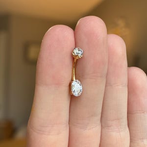 May include: A gold-toned curved barbell navel ring featuring a small round clear gemstone at the top and a larger oval-shaped clear gemstone at the bottom. The metal bar connecting the two stones is smooth and polished, creating a simple jewelry piece.