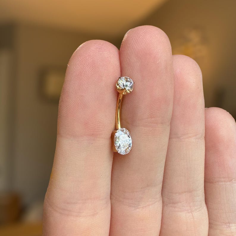May include: A gold-toned curved barbell navel ring featuring a small round clear gemstone at the top and a larger oval-shaped clear gemstone at the bottom. The metal bar connecting the two stones is smooth and polished, creating a simple jewelry piece.
