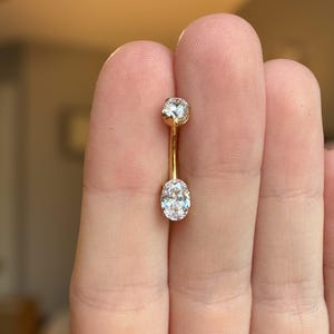 May include: A gold-toned curved barbell body jewelry piece featuring a small round clear gemstone at the top and a larger oval-shaped clear gemstone at the bottom. The metal bar connects the two stones, creating a vertical design for piercing.