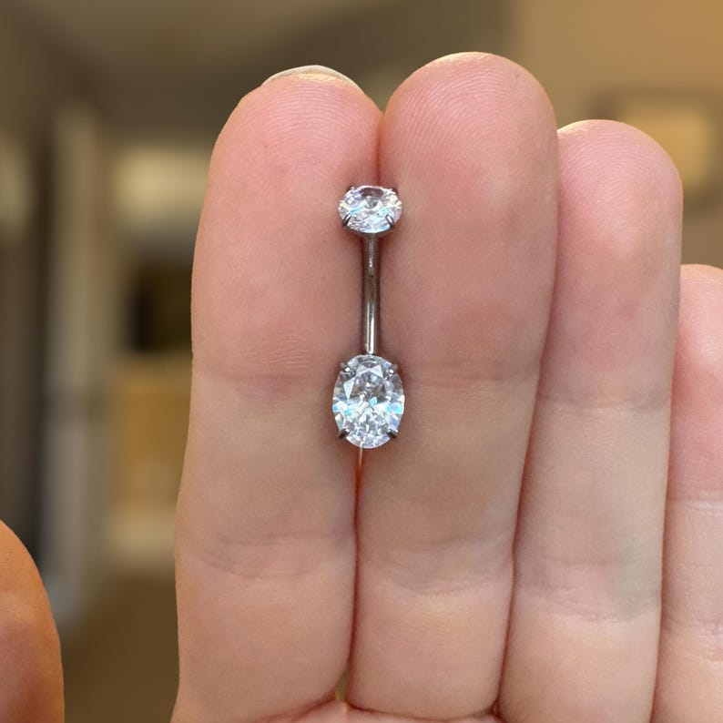 May include: A silver-toned navel barbell featuring a small round clear gemstone at the top and a larger oval-shaped clear gemstone at the bottom. The metal bar connects the two prong-set stones, creating a classic piece of body jewelry.