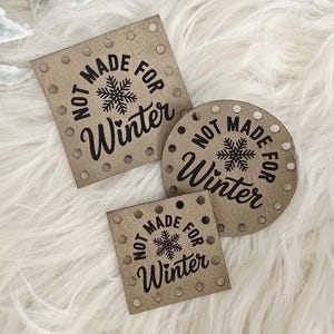 May include: Three brown tags with the text "NOT MADE FOR Winter" and a snowflake graphic. The tags are in square, round, and smaller square shapes. The tags have small holes around the edges. The background is white and fluffy.