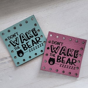 May include: Two square leather patches with the text "Don't Wake the Bear...zzzzzzz" in black. One patch is light blue and the other is pink.