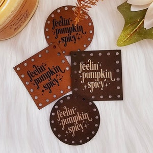 May include: Four brown leather patches with the text "feelin' pumpkin +spicy" in a white font. The patches are in different shapes, including a circle, a square, and two rectangles.