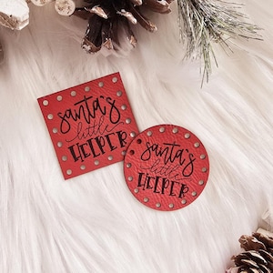 May include: Two red leather ornaments with the text "Santa's little helper" in black lettering. The ornaments are on a white fluffy surface with pine cones.