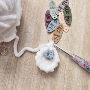May include: A close-up of a crochet project in progress, featuring a ball of white yarn, a crochet hook, and a small crocheted circle. Several small tags with the text "Laser Llama BTQ" and "made in IDAHO" are scattered around the project.