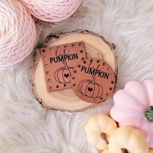 May include: Two brown leather patches with the text "lil pumpkin" and a small pumpkin design. The patches are on a wooden slice with pink yarn balls and miniature pumpkins in the background.