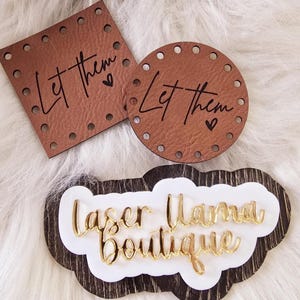 May include: Three brown leather labels with the text "Let them" and a heart symbol. A wooden sign with a white background and gold lettering that reads "Laser Mama Boutique".