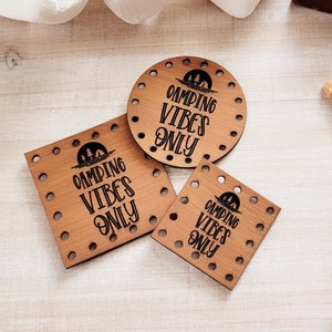 May include: Three wooden signs with the text "Camping Vibes Only" in black lettering. The signs are different sizes and shapes, with a round sign, a square sign, and a smaller rectangular sign. The signs are on a light wood background.