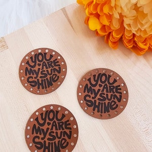 May include: Three brown leather round patches with holes around the edges. Each patch has the text "You are my sun shine" written in black.