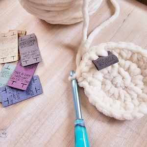 May include: A close-up of a white crocheted  round rug with a brown leather tag that reads "Laser Llama Boutique".  The tag is attached to the rug with a white yarn loop.  There is a turquoise crochet hook and several brown leather tags with the text "Laser Llama Boutique" and "Made in Idaho, Wash Warm, Lay Flat to Dry" on a wooden surface.