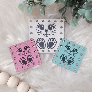May include: Three square felt pieces with a bunny face design. The bunny faces are black and the felt pieces are white, pink, and light blue.