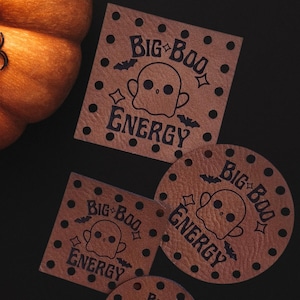 May include: An overhead shot features a large orange pumpkin with a black spider decoration, and two smaller carved pumpkins. Several brown leather coasters with the text "Big Boo Energy" and a ghost graphic are scattered around.