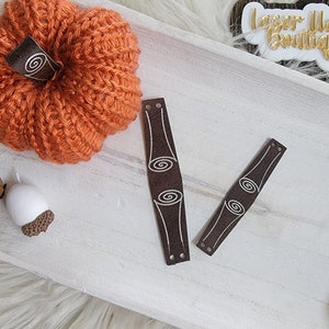 May include: Two brown leather wrist cuffs with a white swirl design. The cuffs are on a white wooden surface.