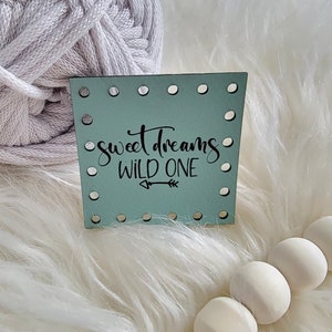 May include: A light blue wooden sign with holes around the edges and the text "sweet dreams wild one" with a small arrow pointing to the right. The sign is sitting on a white fluffy surface.