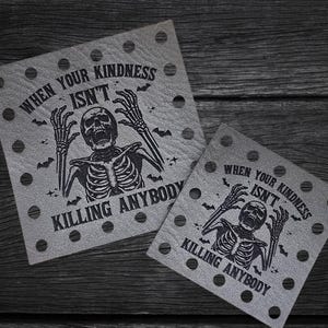 May include: Two gray square coasters with a black skeleton graphic and the text "WHEN YOUR KINDNESS ISN'T KILLING ANYBODY." Each coaster has a series of small holes around the edges. The coasters are on a dark wood surface.