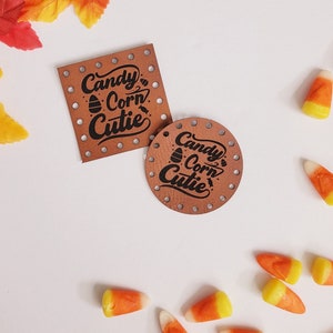 May include: Two brown leather tags with the text "Candy Corn Cutie" in black lettering. The tags have holes around the edges and are surrounded by candy corn.