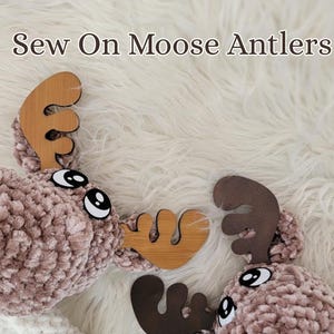 May include: Two plush moose heads with wooden antlers are displayed on a white, fluffy surface. The moose heads are a soft, muted brown color, with large, cartoonish eyes. The text "Sew On Moose Antlers" is at the top.