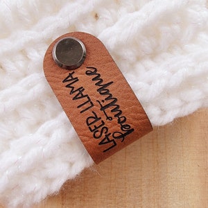 May include: A brown leather tag with the text "LASER LLAMA Boutique" in black script. The tag is attached to a white knitted item with a silver metal rivet. The tag is rectangular with rounded edges.
