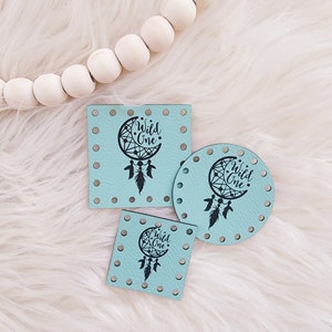 May include: Three turquoise blue leather patches featuring a black dreamcatcher design and the text "Wild One". The patches are in different shapes and sizes: a square, a circle, and a smaller square.