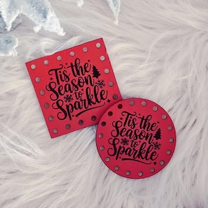 May include: Red square and circular Christmas ornaments with the text "Tis the Season to Sparkle" and a small Christmas tree. The ornaments are on a white, fluffy surface, with decorative frosted leaves and other holiday decorations.