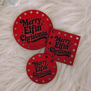 May include: Three red Christmas ornaments with the text "Merry Elfin' Christmas". One is round, one is square, and one is a smaller round shape. The ornaments have a dotted border and are on a white, fluffy surface.