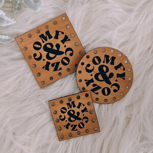 May include: Three wooden coasters with the words "COMFY & COZY" in black lettering. One coaster is square, one is round, and one is smaller and square. The coasters are on a white, fluffy surface, with decorative pinecones and branches.