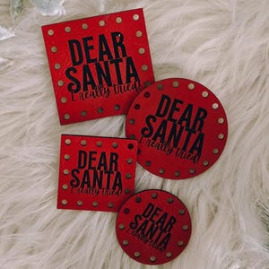 May include: Four red Christmas gift tags with the text "DEAR SANTA I really tried" in black. Two tags are square, and two are round. The tags are on a white fluffy surface, with decorative pine cones and frosted leaves.