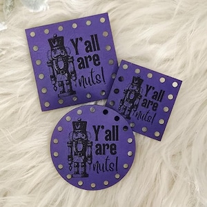May include: Three purple wooden ornaments with the text "Y'all are Nuts!" and a nutcracker design. The ornaments are in square and circular shapes, with small silver dots around the edges. The background is a fluffy, white surface.