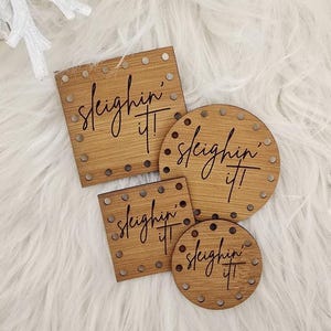May include: Four wooden coasters with the phrase "Sleighin' It" in a script font. Two are square, and two are round, all with small metal studs around the edges. The coasters are on a white, fluffy surface, surrounded by pinecones and decorative elements.