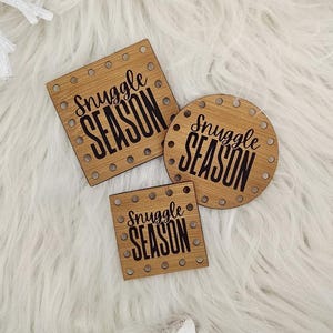 May include: Three wooden coasters with the text "Snuggle Season" in black lettering. One coaster is square, one is round, and one is rectangular. The coasters are surrounded by white decorative elements, including a snowflake, star, and pinecones.