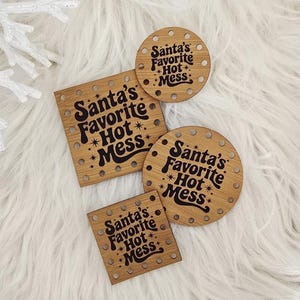 May include: Four wooden coasters with the text "Santa's Favorite Hot Mess" in a black, vintage font. Two are round, and two are square. The coasters are on a white, fluffy surface, surrounded by pinecones, white branches, and a snowflake.