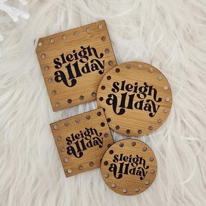 May include: Four wooden coasters with the text "Sleigh All Day" in a black script font. The coasters are in various shapes: square, rectangular, and circular. The coasters are made of light brown wood and are surrounded by a white fluffy surface.