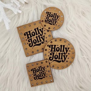 May include: Four wooden coasters with the text "Holly Jolly" in a black, vintage font. The coasters are in various shapes: square, rectangular, and circular. The coasters are light brown with small, dark rivets around the edges. The background is a fluffy, white surface.