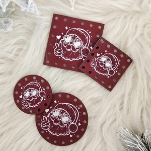 May include: A collection of burgundy-colored Christmas ornaments in various shapes: square and round. Each ornament features a white illustration of Santa Claus with heart-shaped eyes and a peace sign. The ornaments are decorated with small white dots.