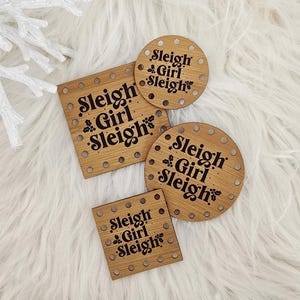 May include: Four wooden coasters with the text "Sleigh Girl Sleigh" in a black font. The coasters are in various shapes: square and round. The coasters are on a white fluffy surface, with pinecones and a snowflake.
