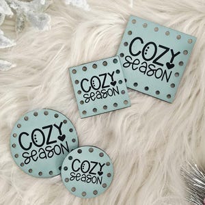 May include: Four light blue wooden tags with the words "COZY season" in black script. The tags are in various shapes: square, rectangular, and circular. Each tag has small, evenly spaced holes around the edges. The tags are on a white, fluffy surface.