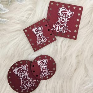 May include: A collection of burgundy-colored coasters and squares with a white illustration of a dog wearing a Santa hat. The coasters are round and the squares have dotted edges. The items are arranged on a white, fluffy surface.