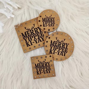 May include: Wooden Christmas ornaments in various shapes, including circles and squares, with the text "MERRY YIPPEE KI-YAY" and a reindeer silhouette. The ornaments are arranged on a white, fluffy surface, surrounded by pine cones and a white star.