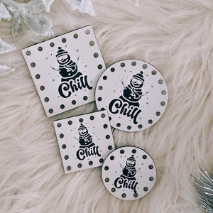 May include: Four wooden coasters with a snowman design and the word "Chill" in black. Two coasters are round, and two are square, all with a white background and black polka dots. The coasters are arranged on a fluffy, white surface.