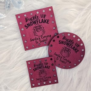 May include: Three red Christmas ornaments with the text "Buckle Up Snowflake" and "Santa's Coming to Town!" in black. The ornaments are square, round, and small square, each with small holes around the edges. The ornaments are on a white, fluffy surface.
